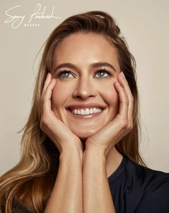 Close-up portrait of smiling model Hailey Carlson with hands framing her face against a neutral beige studio background.