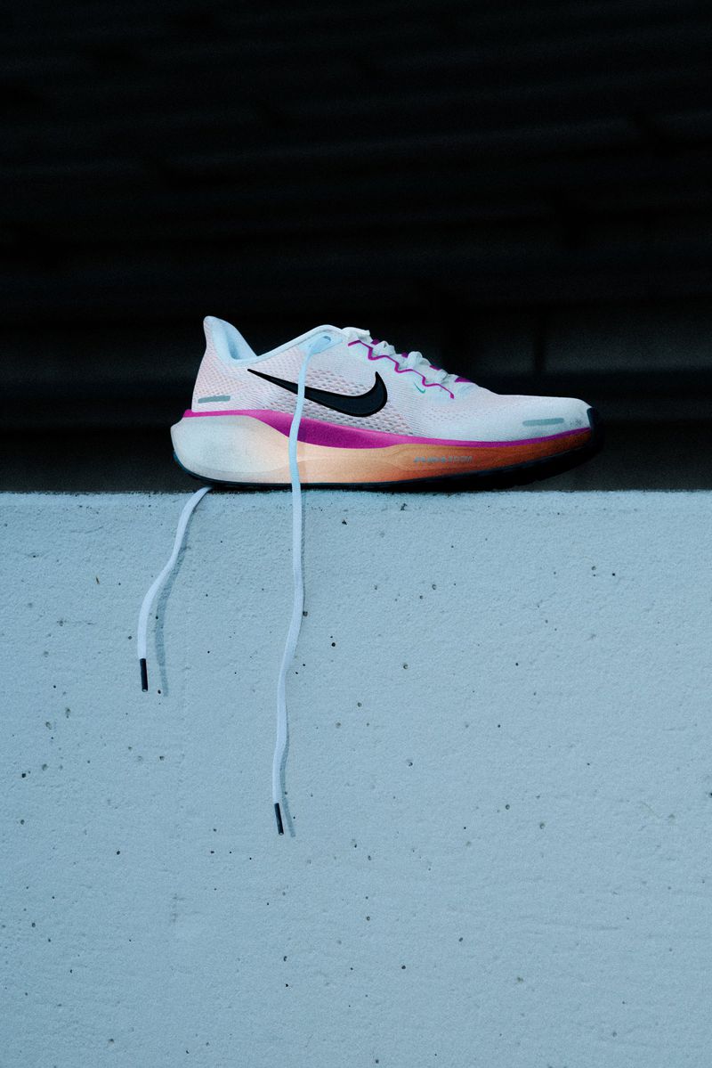 Close-up of a single white, pink, and orange Nike running shoe on a gray concrete ledge with a dark gray background for athletic wear.