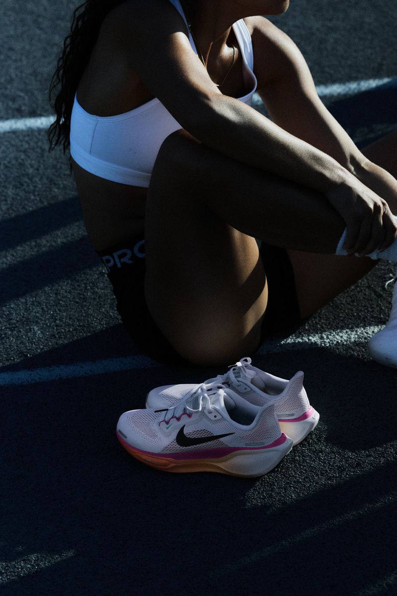Female athlete in sports bra and shorts sitting trackside with white Nike running shoes on dark asphalt, sunlight and shadows.