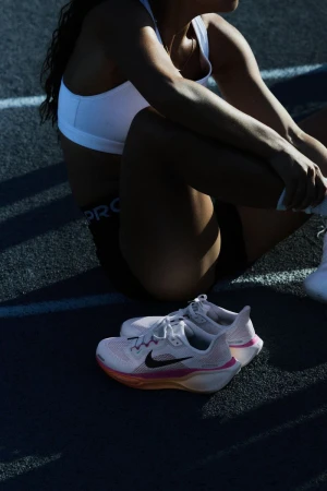 Female athlete in sports bra and shorts sitting trackside with white Nike running shoes on dark asphalt, sunlight and shadows.
