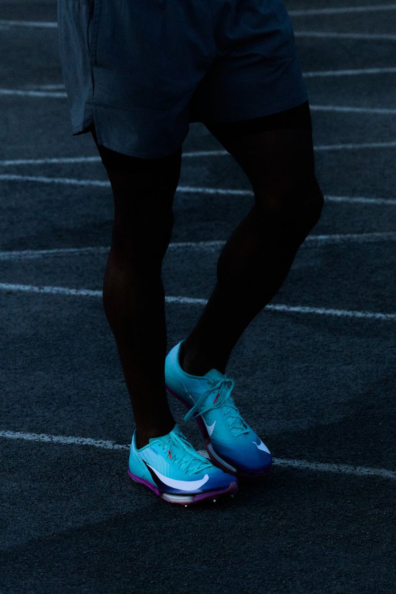 Close-up on an athlete's legs and aqua and purple running shoes, poised on a dark running track, ready for a race.