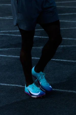 Close-up on an athlete's legs and aqua and purple running shoes, poised on a dark running track, ready for a race.