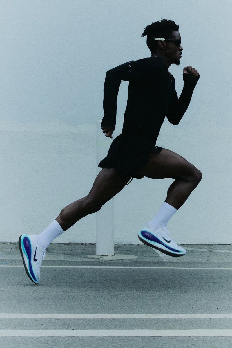 Athletic man running outdoors on an asphalt surface wearing dark workout gear and white running shoes with blue accents against a neutral wall.