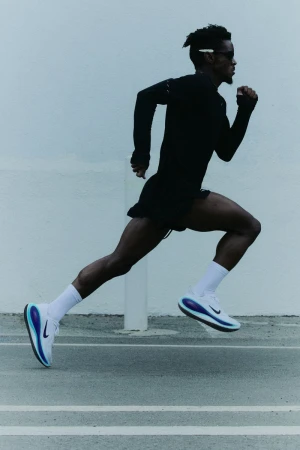 Athletic man running outdoors on an asphalt surface wearing dark workout gear and white running shoes with blue accents against a neutral wall.