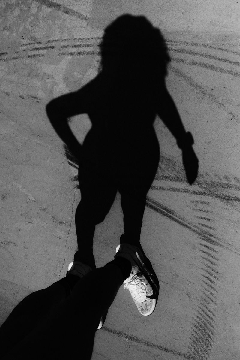 Monochrome image of a woman's silhouette with curly hair, visible legs, and a wrist accessory, alongside a person's leg and Nike shoe.