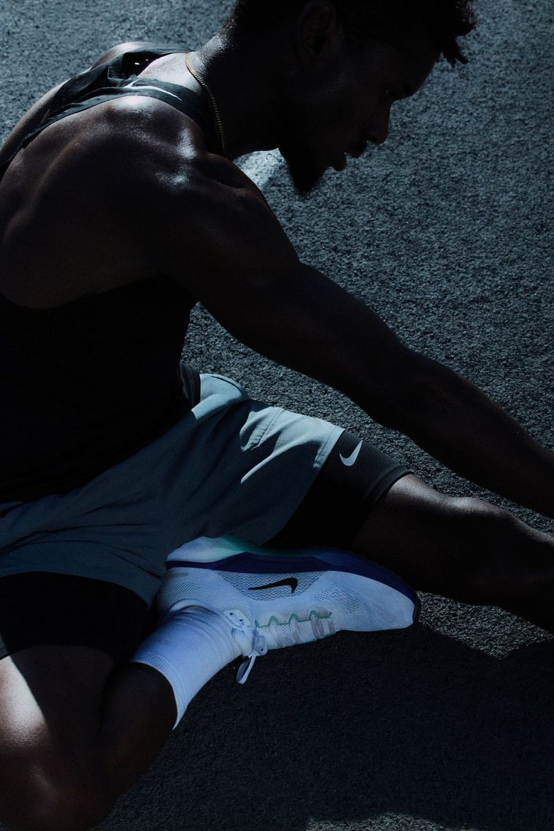 Man stretching on asphalt, wearing a black tank top, grey shorts, white socks and shoes, captured in low lighting with visible sweat.