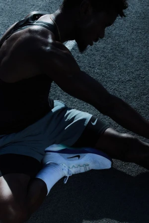Man stretching on asphalt, wearing a black tank top, grey shorts, white socks and shoes, captured in low lighting with visible sweat.