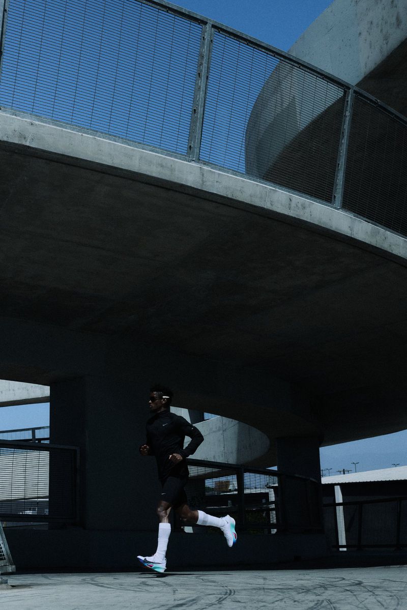 Adult male athlete running outdoors in sportswear with a concrete bridge overhead and a blue sky visible in the background.