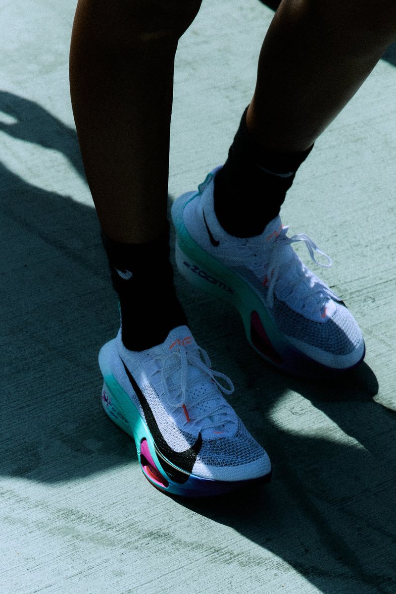 Close-up of person wearing white running shoes with teal and black Nike swoosh logo and black socks on pavement with harsh shadows.
