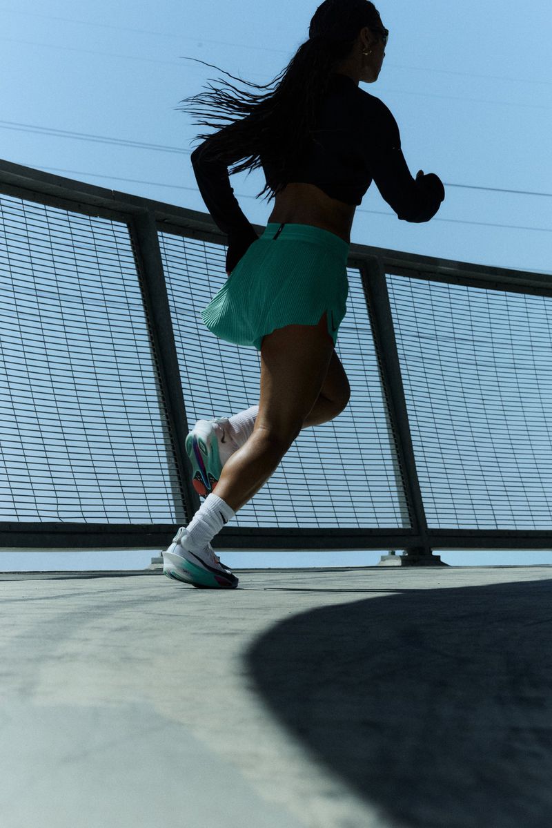 Athletic woman in sport clothing runs on a bridge in a bright, sunny outdoor setting with a metal fence under clear blue sky.