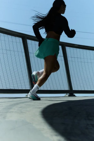 Athletic woman in sport clothing runs on a bridge in a bright, sunny outdoor setting with a metal fence under clear blue sky.