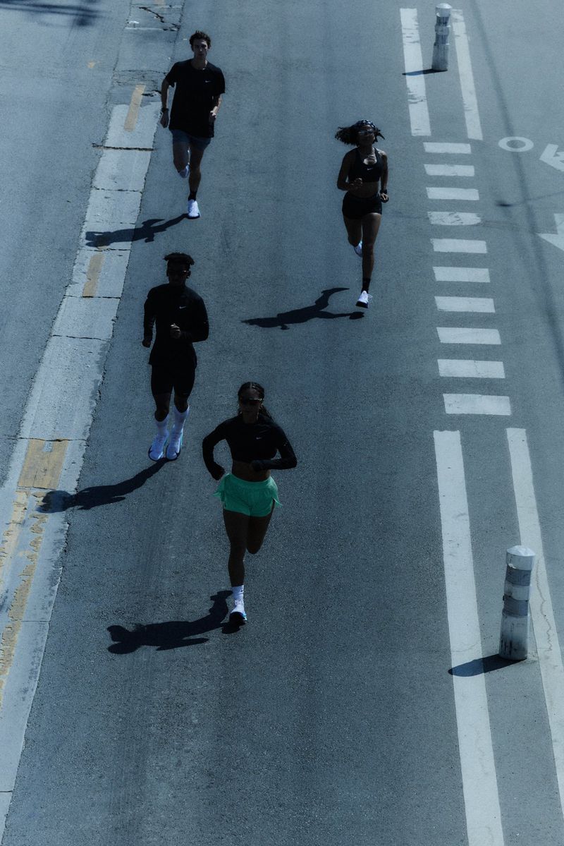 Overhead shot of four diverse adults jogging on an asphalt road with painted markings, under bright sunlight, casting long shadows.