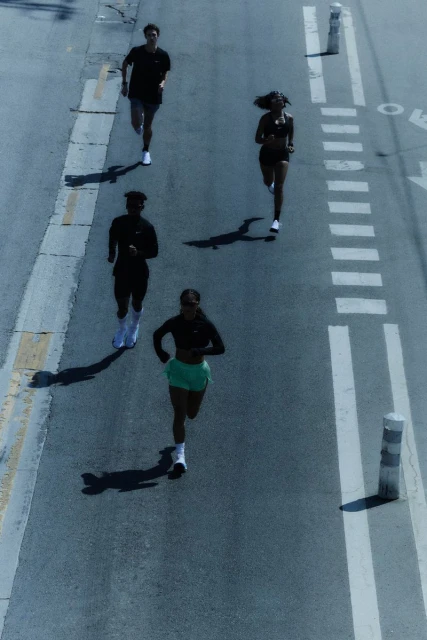 Overhead shot of four diverse adults jogging on an asphalt road with painted markings, under bright sunlight, casting long shadows.