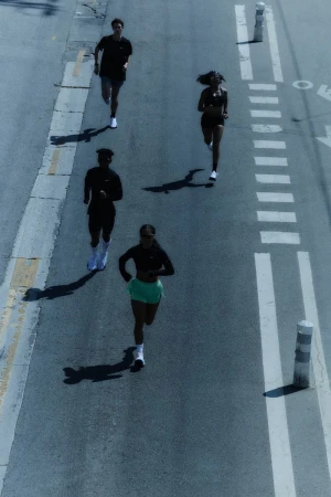 Overhead shot of four diverse adults jogging on an asphalt road with painted markings, under bright sunlight, casting long shadows.