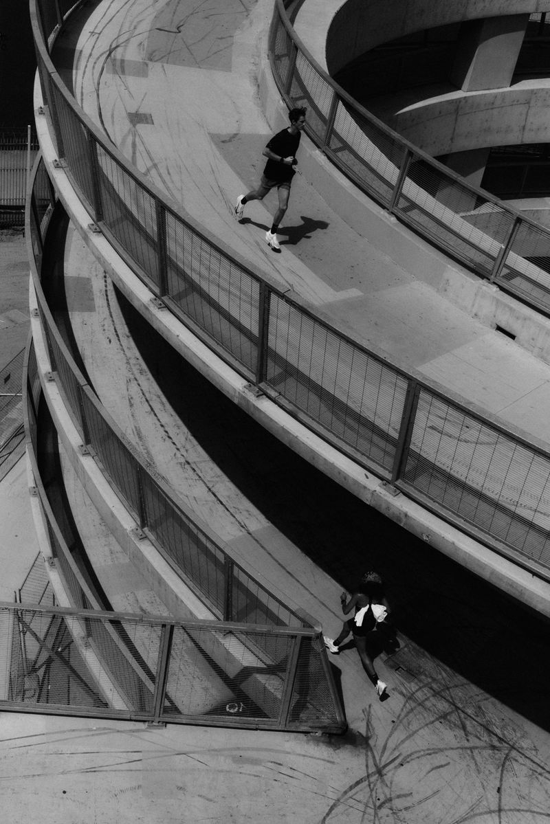 A man is running on a concrete circular ramp with metal railing while another person is standing on the bottom level in monochrome.