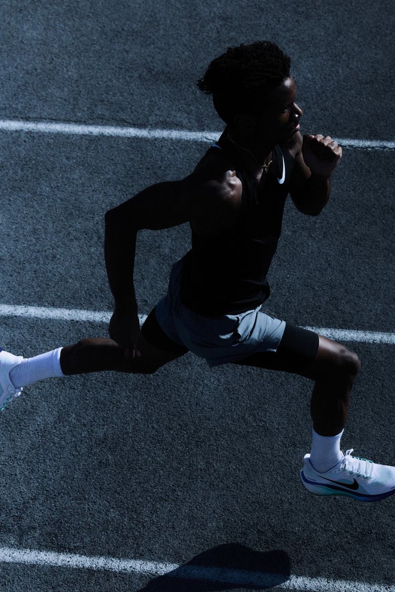 Action shot of a male athlete with dark skin and curly hair in mid-stride on a running track under bright lighting, captured from above.
