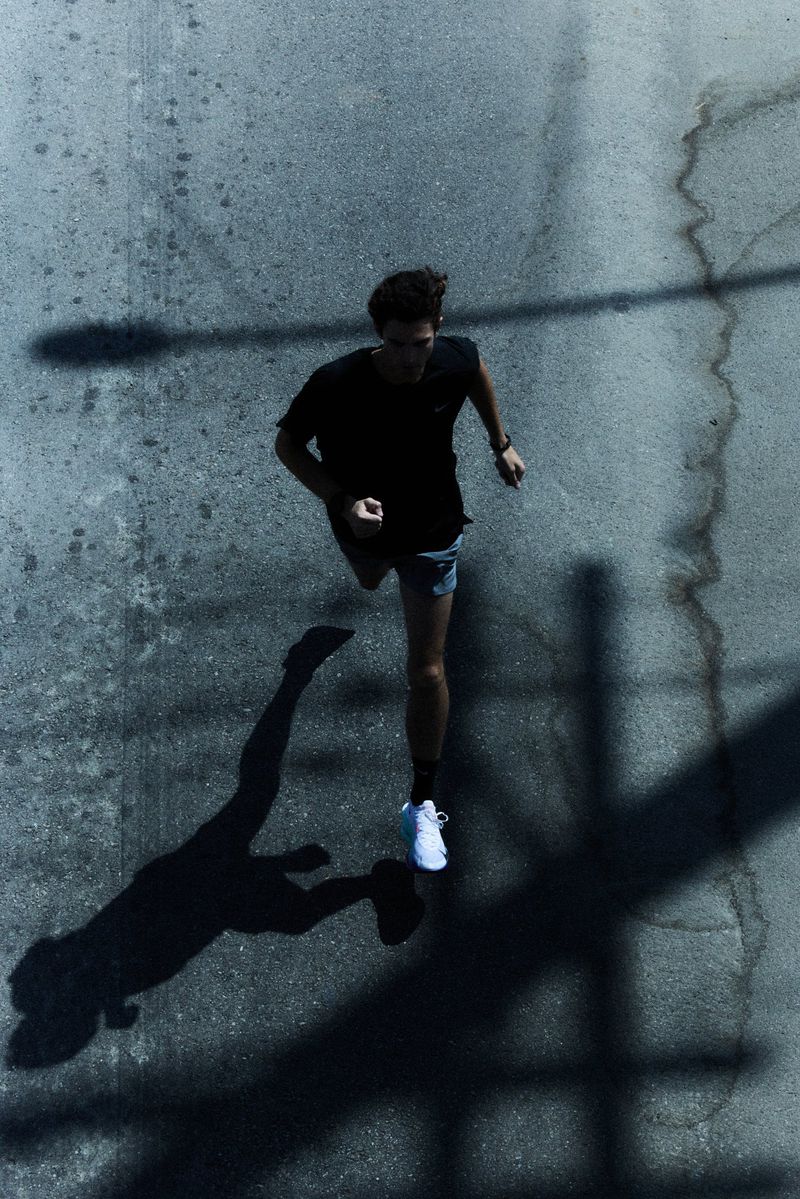 High-angle shot of a young man running on an asphalt road, casting a long shadow. He wears a black t-shirt, shorts, and white sneakers.
