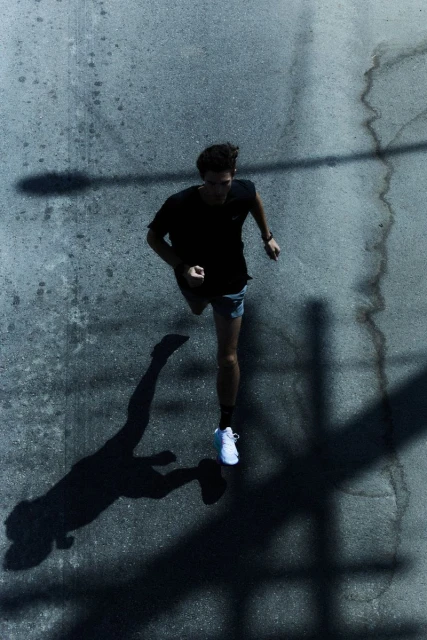 High-angle shot of a young man running on an asphalt road, casting a long shadow. He wears a black t-shirt, shorts, and white sneakers.