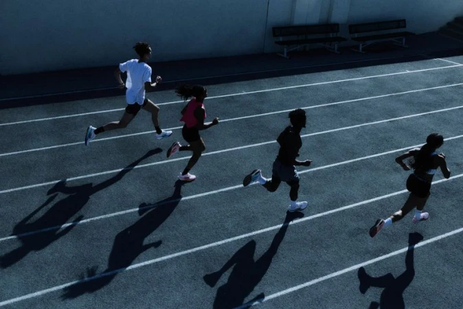 Four runners sprint on a blue track with white lane lines, captured from above, casting long shadows under bright lighting, highlighting athleticism and motion.