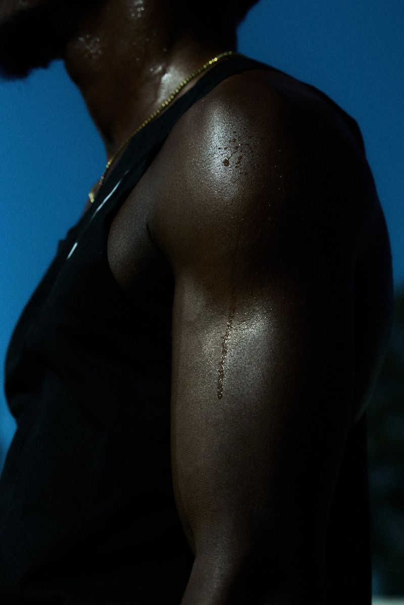 Muscular Black man in a sleeveless top, sweat glistening on his arm against a gradient blue sky. Close-up shot emphasizes fitness and athleticism.
