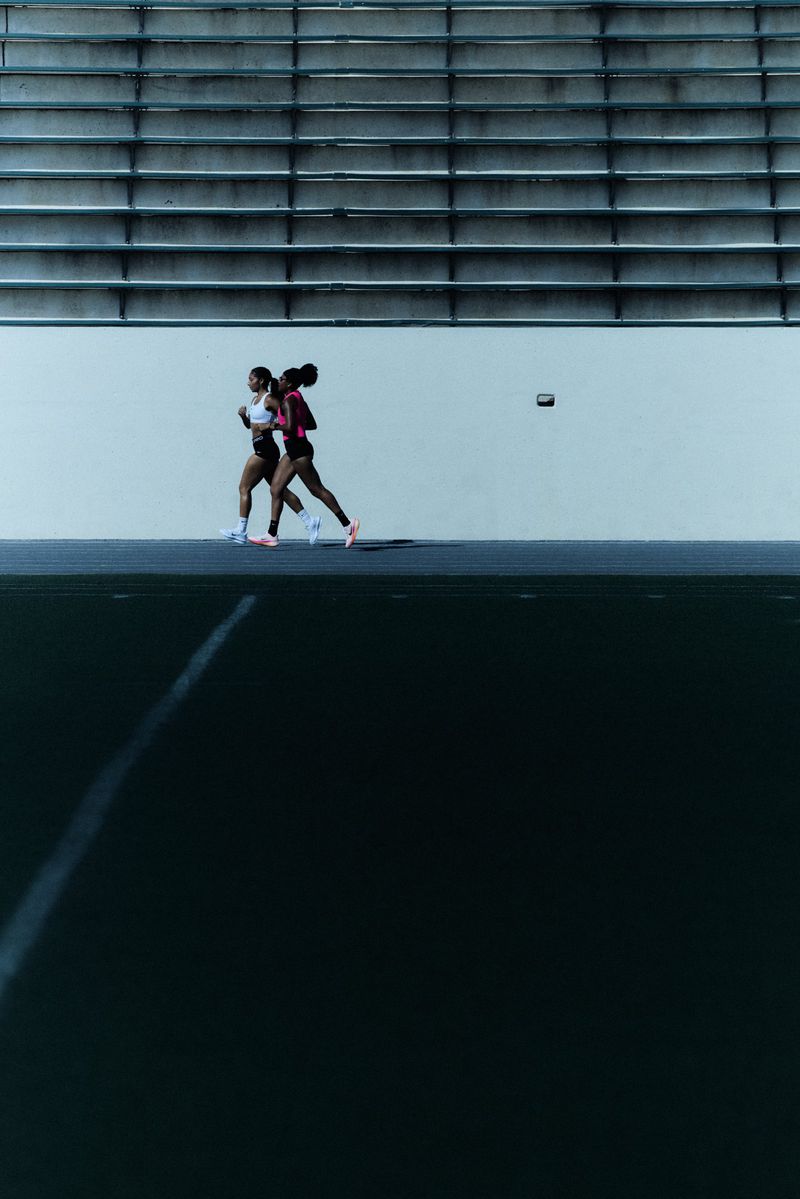 Two African-American women runners run side-by-side on a track in athletic wear with stadium seating in the background.