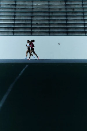 Two African-American women runners run side-by-side on a track in athletic wear with stadium seating in the background.