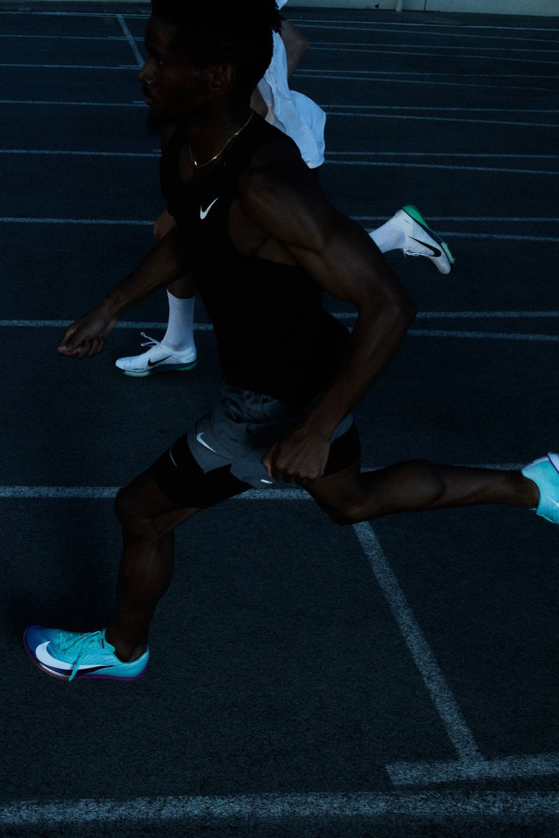 Athletic man sprints on a blue running track wearing Nike apparel, racing against another runner in an outdoor setting during the day.