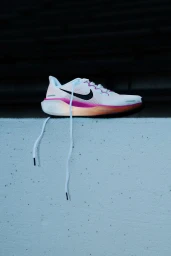 A white Nike running shoe with a pink and orange gradient sole rests on a light concrete ledge with laces hanging down.