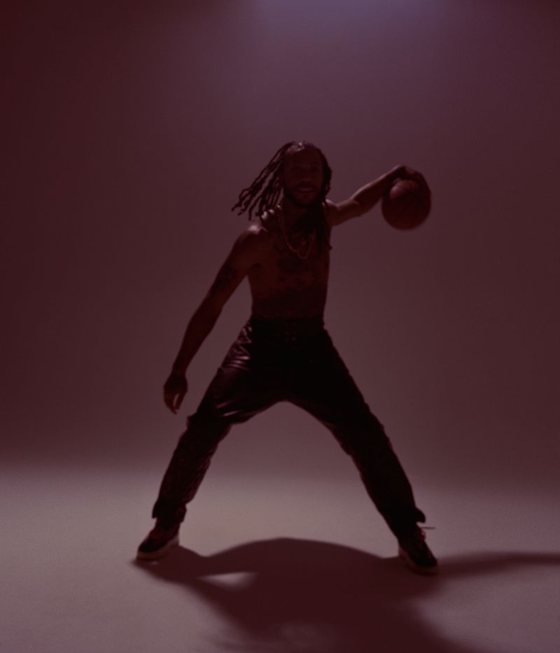 Athletic adult male with dreadlocks, shirtless, in leather pants, is holding a basketball in a studio with a red background.