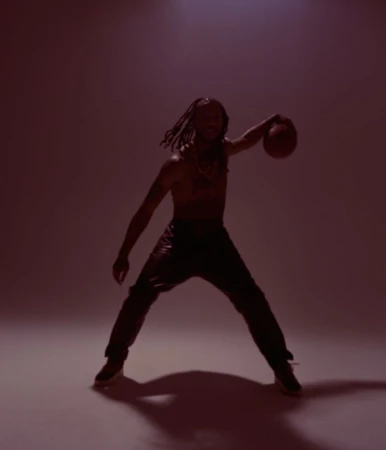 Athletic adult male with dreadlocks, shirtless, in leather pants, is holding a basketball in a studio with a red background.
