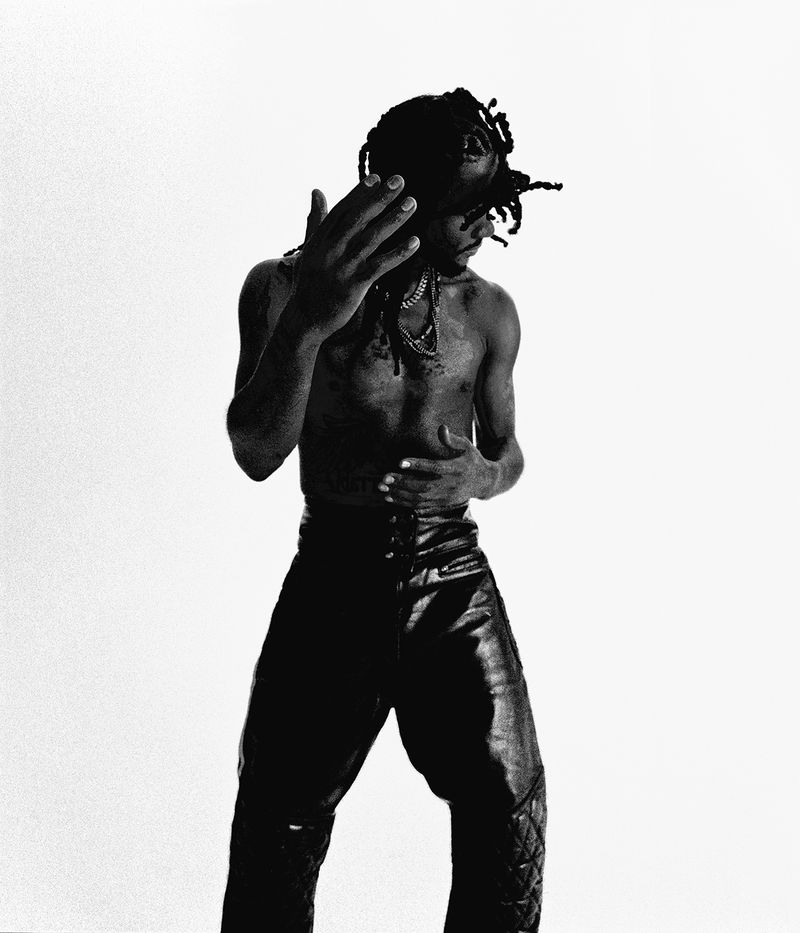 Black and white portrait of a shirtless Black man with dreadlocks wearing shiny black leather pants posing against a bright white backdrop.