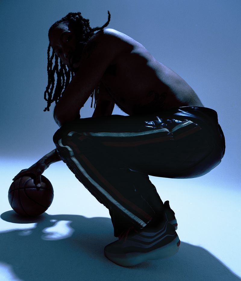 Athletic African American male squats indoors in blue light, shirtless, holding basketball in photo studio. He wears striped pants.