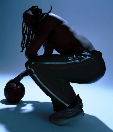 Athletic African American male squats indoors in blue light, shirtless, holding basketball in photo studio. He wears striped pants.