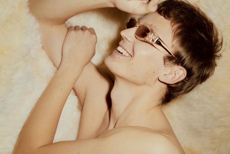 Close-up studio portrait of a young adult male with sunglasses lying on fluffy white surface, smiling and looking up.