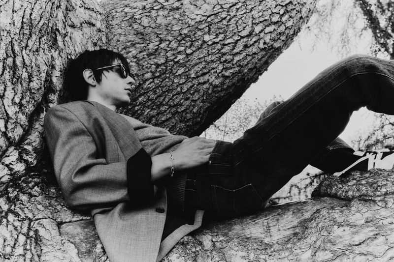 Monochrome portrait of a man in a blazer and sunglasses reclining in a tree, with visible bark texture.