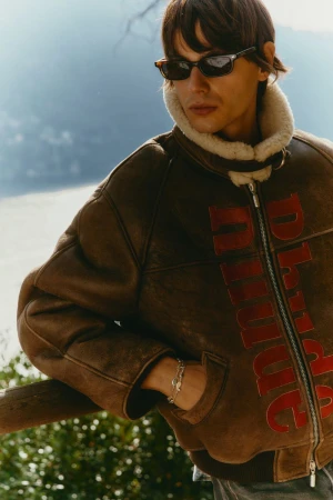 Man in brown shearling jacket and sunglasses poses outside with blurred natural background, showcasing fall or winter fashion apparel.