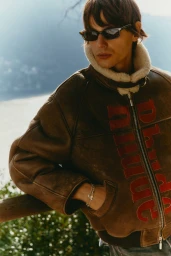 Model wearing a brown shearling-collar leather jacket with red lettering outdoors near a body of water