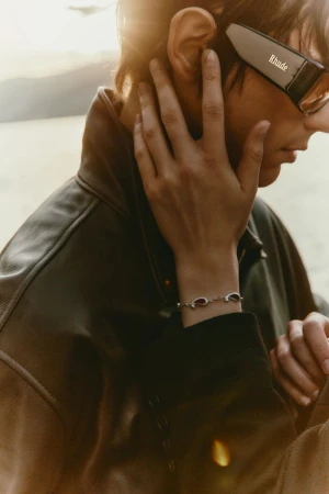 Close-up profile of a model wearing Rhude sunglasses and a leather jacket as a hand wearing a bracelet touches his face.