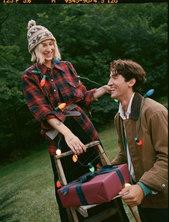 Couple exchanges gifts outdoors; woman in hat and plaid shirt holds ladder with present; man in jacket smiles wearing lights.