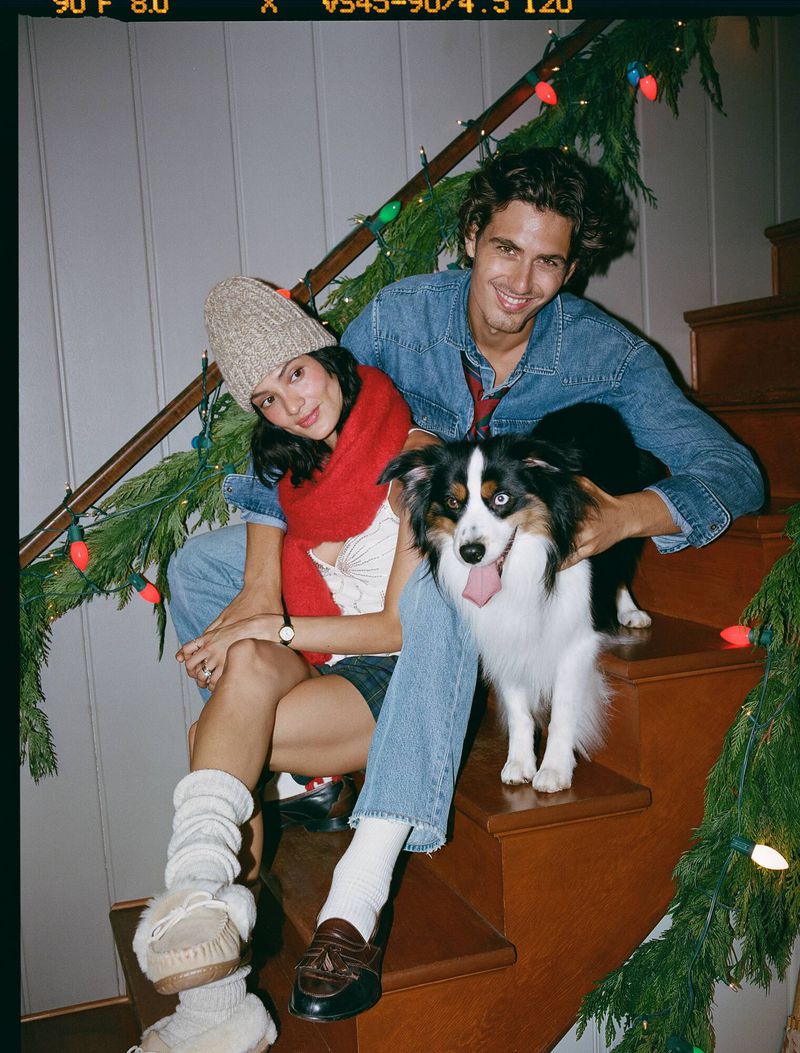 Young couple and a dog sitting on stairs decorated with holiday lights and garlands in a cheerful, casual holiday fashion setting.