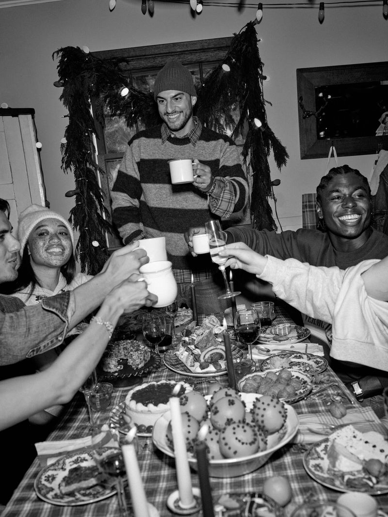 Group of young adults toasting with mugs and glasses at a dining table laden with food, in a festive indoor gathering, monochrome.