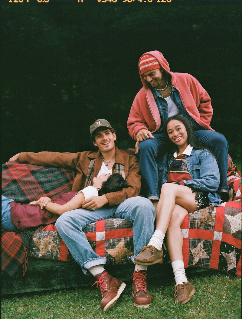 Four young adults in casual clothes are outdoors on a vintage quilt-covered sofa, smiling and relaxed on a sunny day, surrounded by dark foliage.