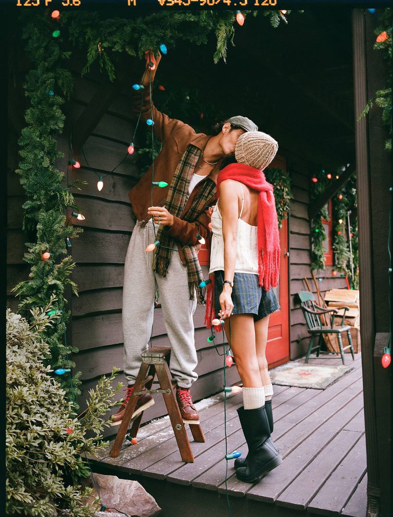 Couple kissing on cabin porch, decorating Christmas lights. Woman in shorts, hat, boots; man on ladder in sweats & brown jacket.