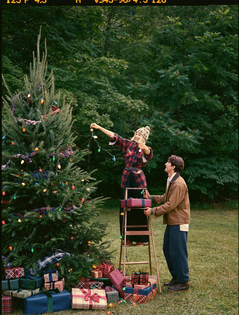 Woman on ladder decorates Christmas tree as man holds ladder in outdoor setting with wrapped presents and natural green backdrop.
