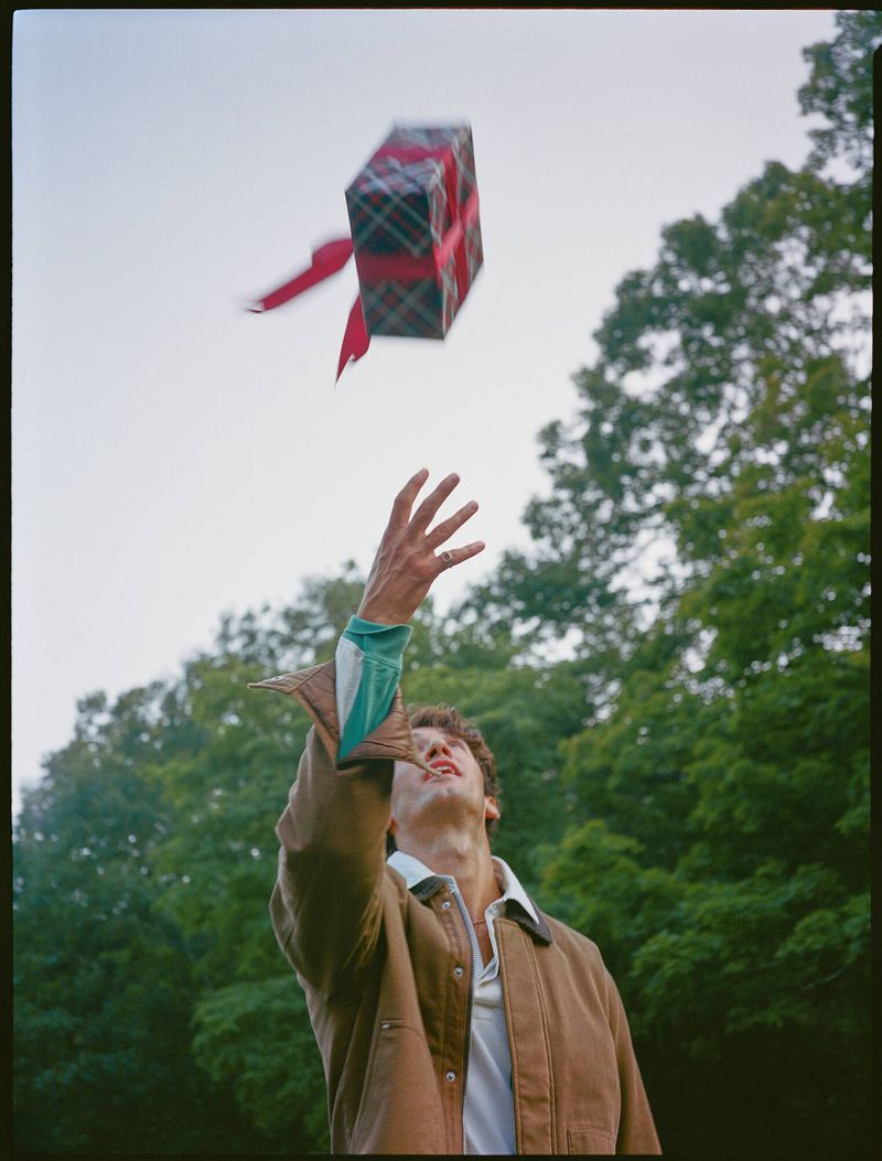 Young man in a brown jacket reaches for a present box floating in the air outdoors, trees and sky are visible in the background.