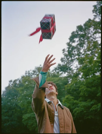 Young man in a brown jacket reaches for a present box floating in the air outdoors, trees and sky are visible in the background.