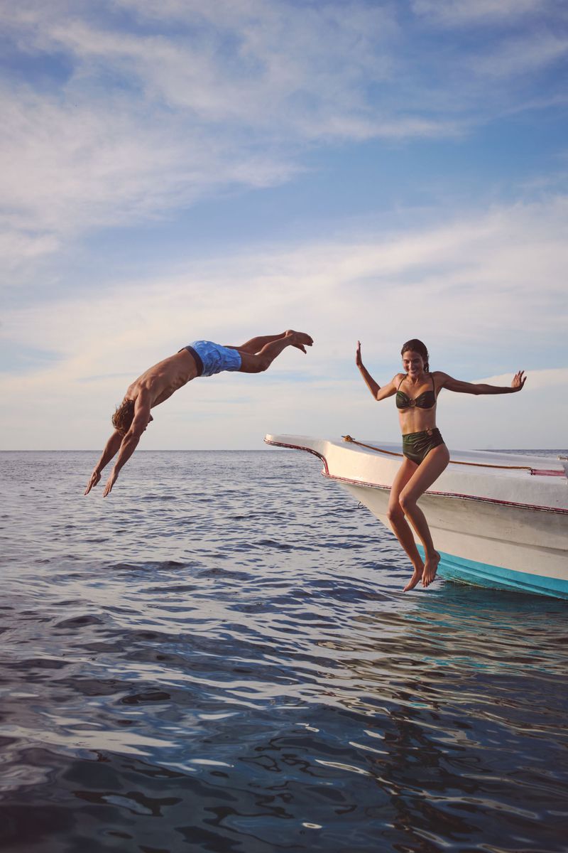 Adult male dives into ocean water from boat near adult female jumping, under blue sky with clouds, sunny day, summer vacation.