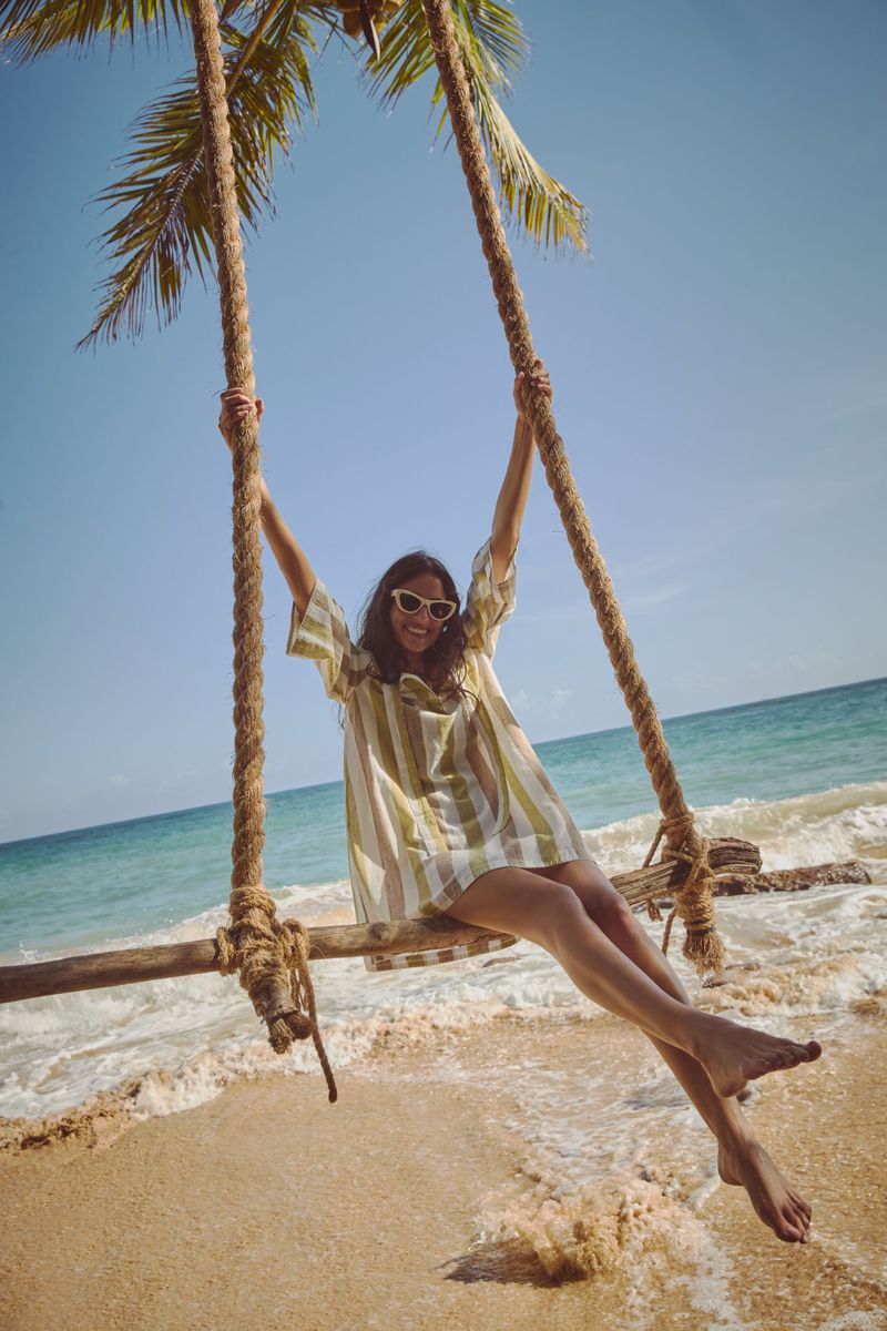 Woman in summer dress on a rope swing hanging from a palm tree on a sandy beach with turquoise ocean and clear blue sky in the background.
