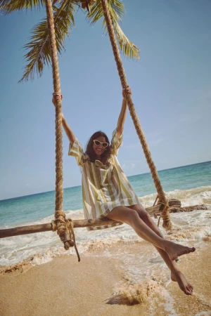 Woman in summer dress on a rope swing hanging from a palm tree on a sandy beach with turquoise ocean and clear blue sky in the background.