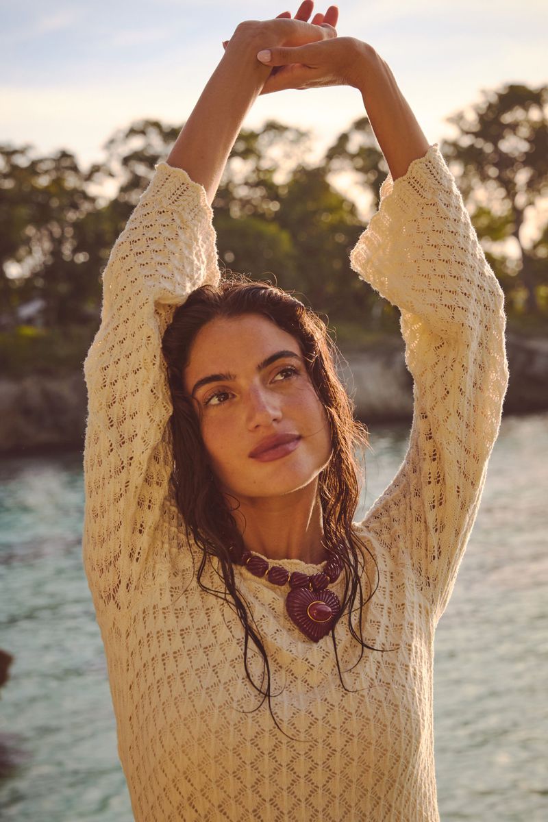 Woman in white dress with arms raised outdoors. She wears a large heart necklace. Trees and sky fill the background.